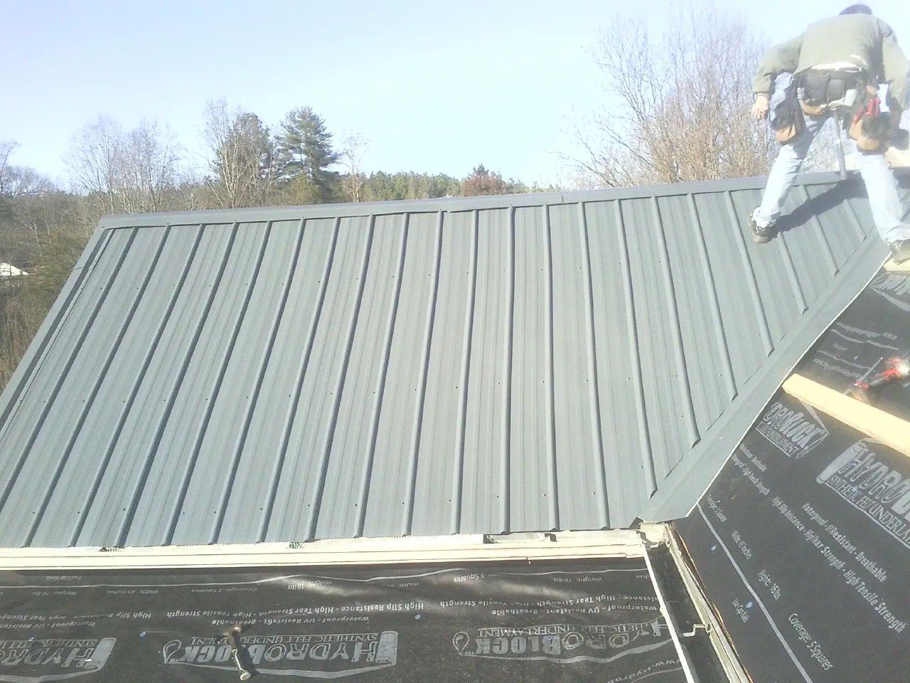 Crew installing dark green standing seam panels over synthetic underlayment in West Salem