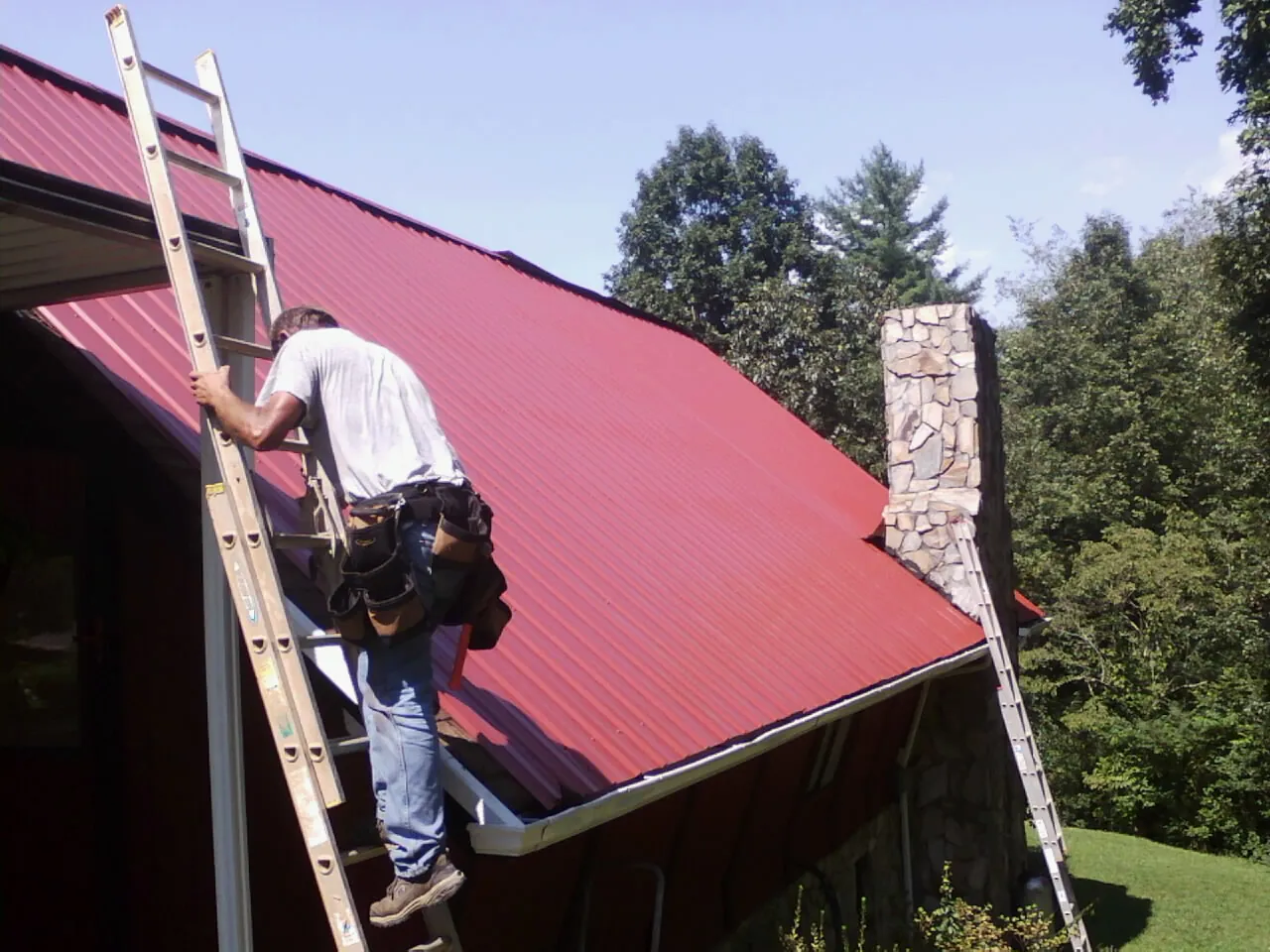 Roofer on a ladder beside a red metal roof and stone chimney — Soffit & Fascia Repair by Roofing Brothers in West Salem